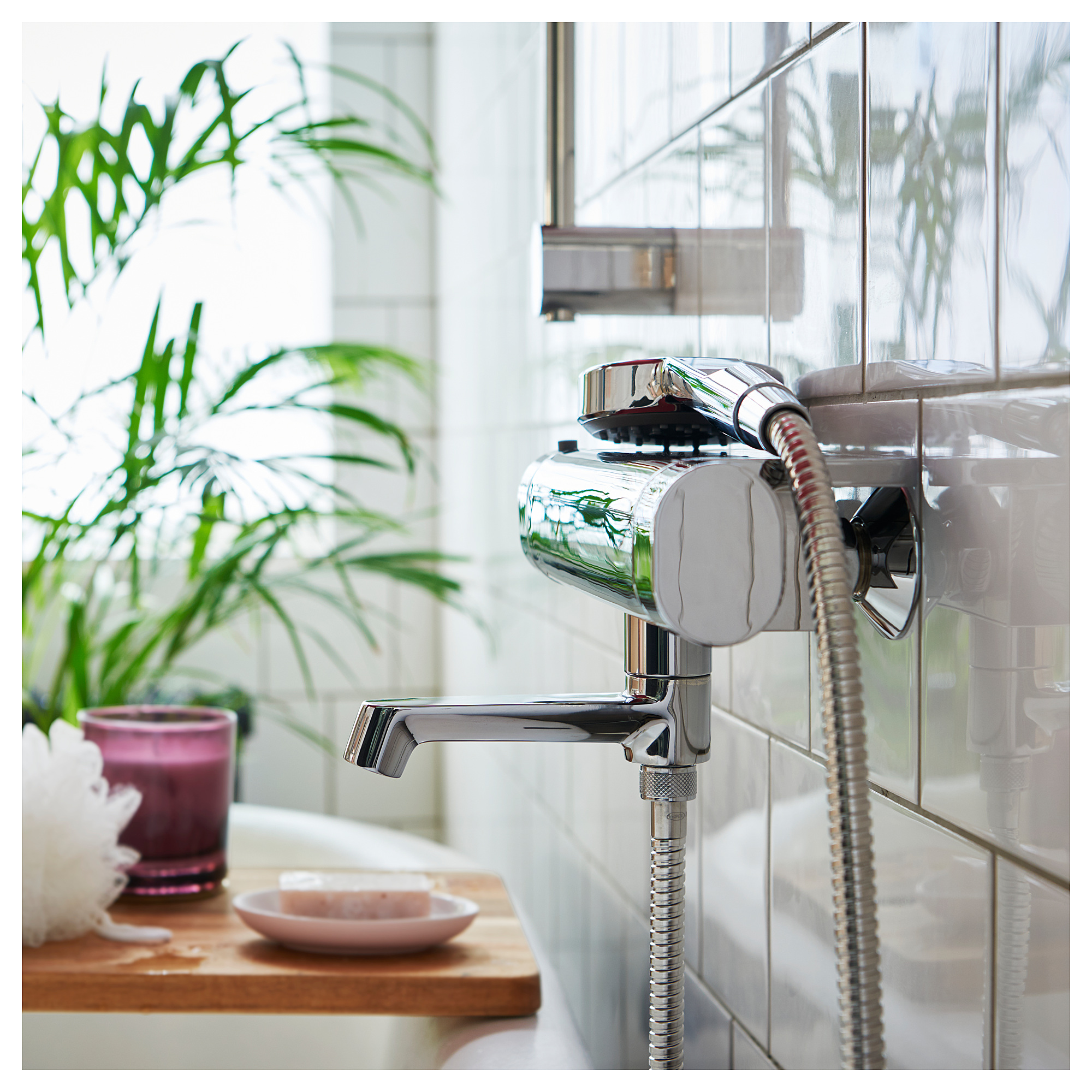 Modern silver-finish bathroom faucet with handheld showerhead, mounted on a white tiled wall beside a green plant in a purple vase on a wooden shelf.