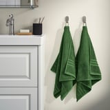 BROKGLIM organic towels in bathroom. Dark green, 100% cotton, folded on silver hooks beside sink. Soft, absorbent, with woven border.