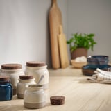 BRUGDHAJ jars, white with cork tops, hold various items on a wooden table by a potted plant, showing size comparison with wooden utensils.