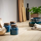 Three blue ceramic jars with cork lids on wooden table, dishes, stacked bowls, bottle, and plant.