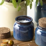 Two blue ceramic jars with cork lids, one filled with salt, on a wooden surface. Fresh herbs in a vase nearby.