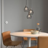 Modern dining area with three hanging BRUNSTA lamps, small, geometric, black metal shades, above a round light wood table with orange chairs.