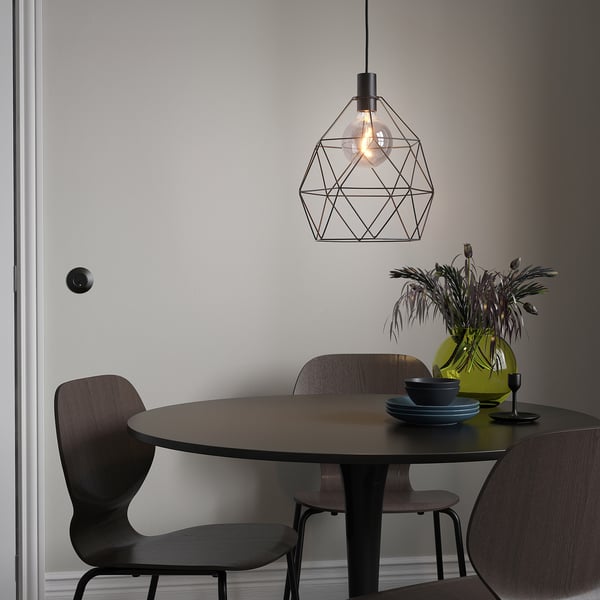 Modern dining setup with BRUNSTA lamp, small dark table, wooden chairs, green vase with flowers, and blue plates.