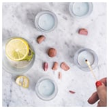 Small candles in blue glass on marble. Lemon slice drink beside. Hand holding lighter. Wood pieces scattered.