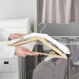 Hand holds whitewood BUMERANG hanger, preserving clothes shape, near washing machine and closet.