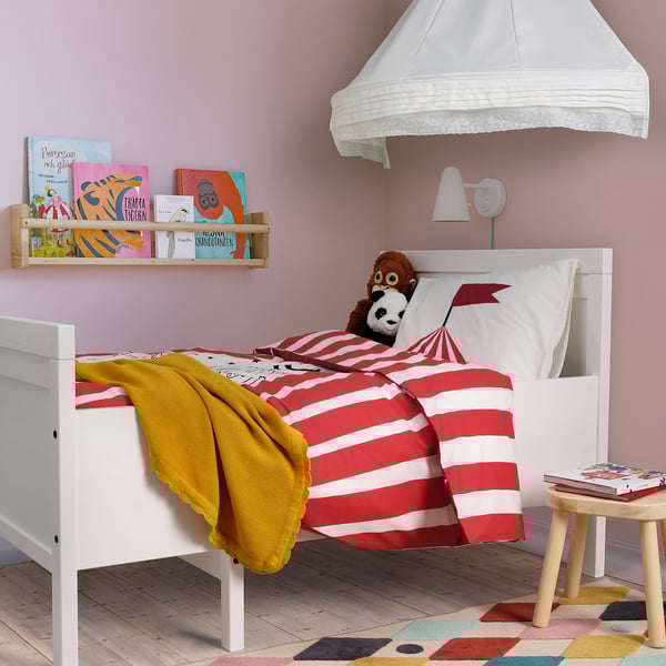 Colorful kids room with BUSENKEL red & white striped bedding, monkey plush, books, yellow throw, wooden stool, pastel rug, white walls & canopy.