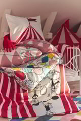 A cozy bedroom featuring a BUSENKEL circus-themed duvet cover in red and white stripes; colorful, playful design with various circus characters.