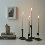 Three lit twisted candles in black holders on a surface, next to books and a wooden house decoration, with a framed picture on the wall behind.