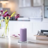 Small lit lavender DAGLIGEN candle sits on glass holder, beside vase with flowers and stacked books on white table.