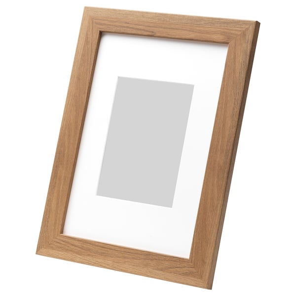 Light brown wooden picture frame, leaning against white wall, rectangular shape, neutral photo mount.