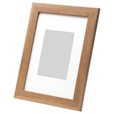 Light brown wooden picture frame, leaning against white wall, rectangular shape, neutral photo mount.