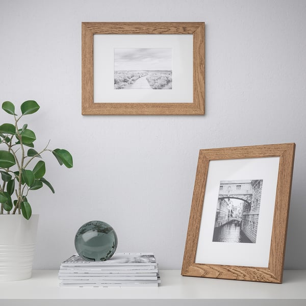 Two wooden picture frames display landscapes; one hangs, the other stands. Beside a plant, a stack of books holds a decorative sphere.