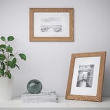 Two wooden picture frames display landscapes; one hangs, the other stands. Beside a plant, a stack of books holds a decorative sphere.