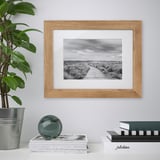 A white shelf displays a wooden picture frame with a black-and-white a wooden path, surrounded by books, a plant, and a decorative sphere.