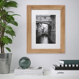 Black and white a venetian bridge in a light wood frame on a white wall. Below it, a stack of books with a silver ornament and a potted plant.