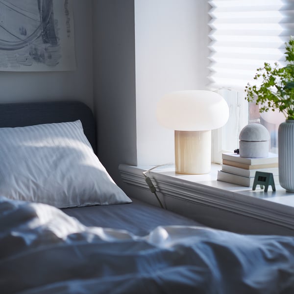 Cozy bedroom with DEJSA white lamp, silk bedding, grey headboard, blue art, and potted plants.