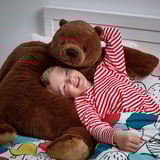 Child in striped pajamas hugs large brown teddy bear on bed.