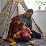 Two people sit inside a gray teepee with a large brown teddy bear, suggested for cuddling and play, suitable from 18 months.
