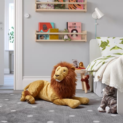 Large yellow lion stuffed toy lays on childs room floor, surrounded by books, small toys, and bed.