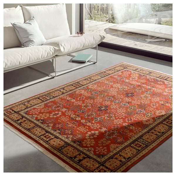 Bright red ornate rug with floral patterns, bordered by intricate designs, lies near white couch and a window.