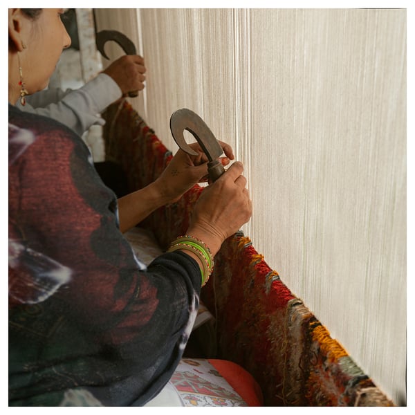 Person weaving fabric on traditional loom, hands detailed, colorful bracelets visible, wearing red and black patterned shirt.