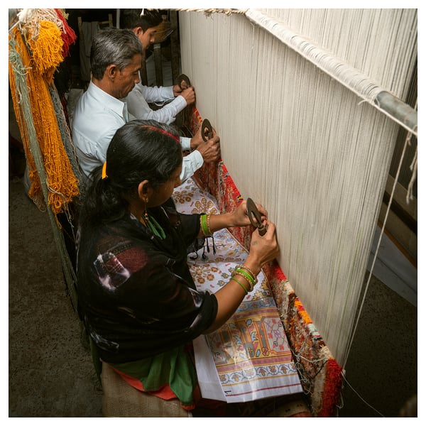 Three people weave fabric on a loom. Traditional tools and colorful threads visible. Man and woman work together, focused on their task.
