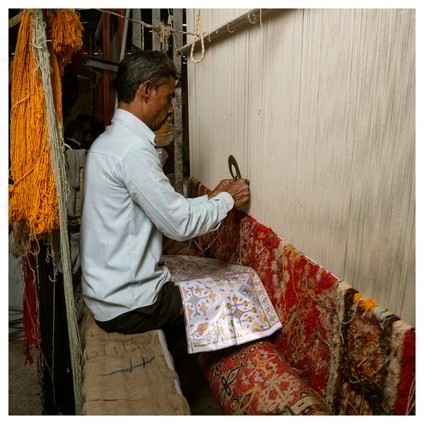 Man weaving colorful patterned rug on a loom. Bright yarns hang on left. Rug displays floral design in red, yellow, and blue.