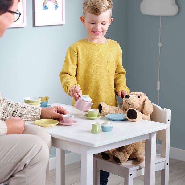 Child pours tea with teddy bear; woman smiles.