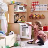 Child playing with DUKTIG toy kitchen. Girl in pink dress pretends to bake, surrounded by toy oven, fruits, and stuffed animals.