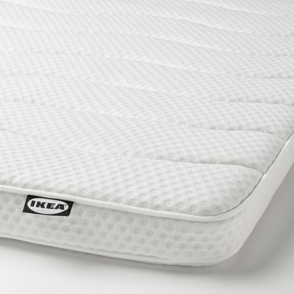 Displays a close-up of an IKEA mattress, showcasing its textured white surface and quilted edges. A black IKEA logo tag is visible on one corner.