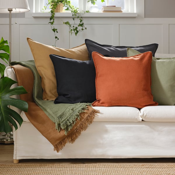 White couch with dark beige, navy, and orange pillows. Green throw blanket, indoor plant, and white lamp nearby.