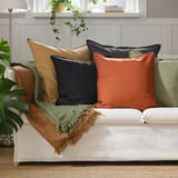 White couch with dark beige, navy, and orange pillows. Green throw blanket, indoor plant, and white lamp nearby.