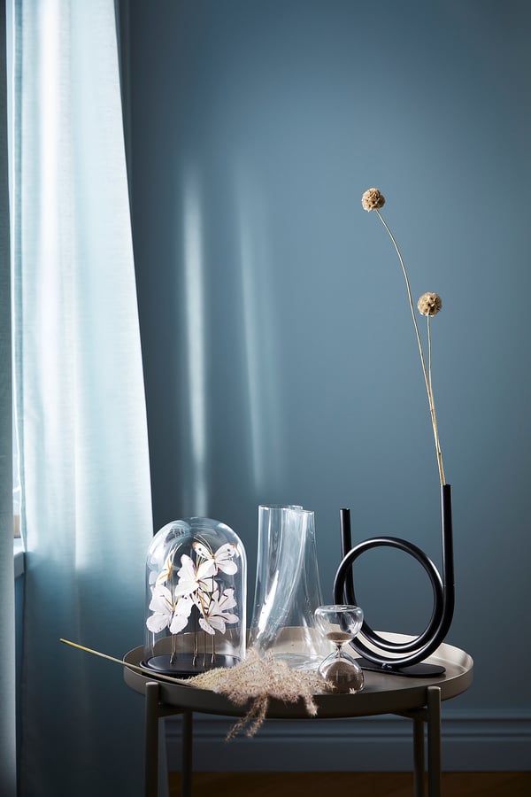 Clear glass hourglass with tinted finish, modern design, stands on metal stand, minimalist decor, blue wall background.