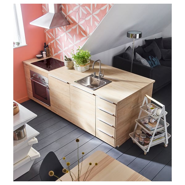 Small, light wood kitchen with sink, oven, and storage. Bright living area with black couch and colorful wall.