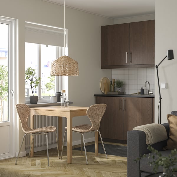 Modern kitchen nook with wooden table, two woven chairs, pendant lamp, & dark wood cabinets. Bright, airy space with large window.