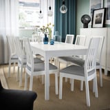 Modern white extendable dining table with six matching chairs, some with grey cushions. Set in a well-lit room with natural decor.