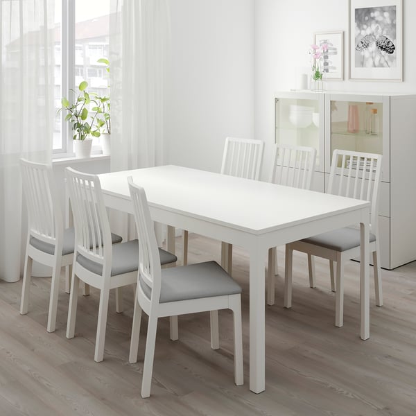 Minimalist white dining table with 6 gray-cushioned chairs, modern vertical-slat backs, set against a window.