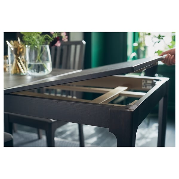 Person opens dark extendable table, stylish chairs visible, green wall backdrop.