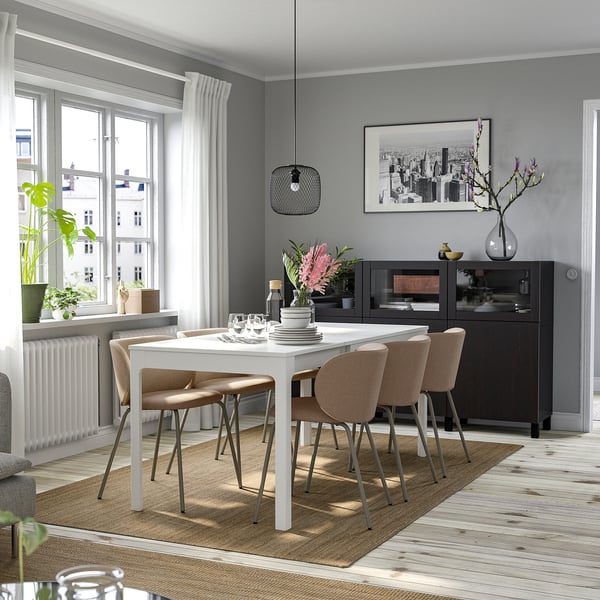 Modern dining room with EKEDALEN extendable white table, four beige chairs, rug, vase, city print, bright, large windows, wooden floor.