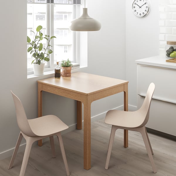 Brown oak dining set with 2 beige chairs near window with plants.