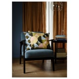 Modern graphic cushions on a grey retro-styled chair with wooden legs, positioned near a small table and a tall window with brown curtains.