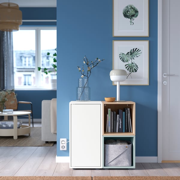 Modern blue room with white EKET storage unit. Vase with flowers, lamp, books, and framed plant art.