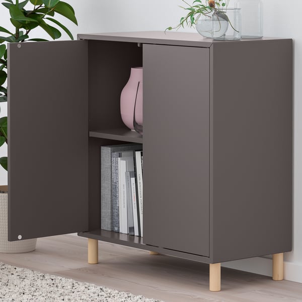 Modern dark cabinet with open doors revealing books and a vase. Mounted on wooden legs, includes decorative items on top.