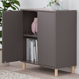 Modern dark cabinet with open doors revealing books and a vase. Mounted on wooden legs, includes decorative items on top.