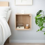 Minimalist wooden wall niche, housing alarm clock, glass, books, and plant, beside bed.