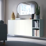 EKET wall cabinet in white with open & closed storage, below mirror, books, cactus, next to teal chair.