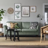 Modern living room with EKHOLMA sofa, green, plush, 3-seater. Wall art, floor lamp, wicker chair, and rug. Clean, minimalist design.