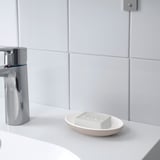White oval soap dish on a bathroom counter, holding a bar of soap, next to a shiny chrome faucet. Part of the EKOLN series.