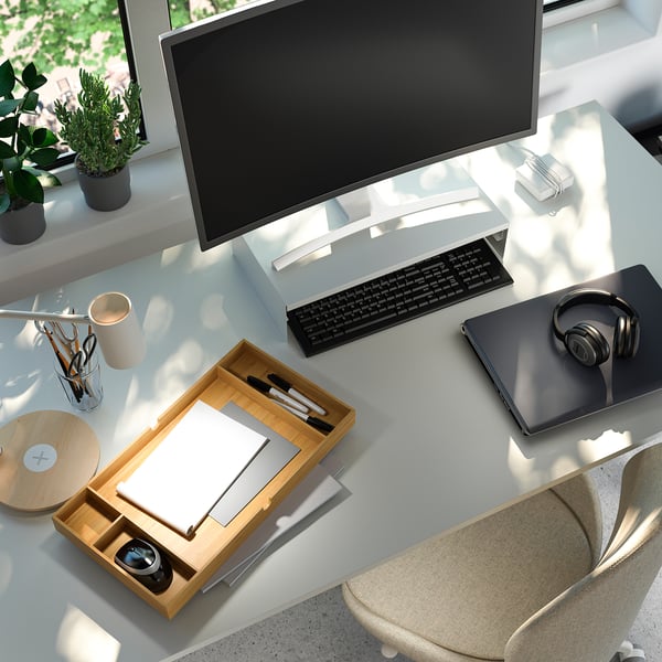 Organized workspace with wooden monitor stand, keyboard, laptop, headphones, and plants.