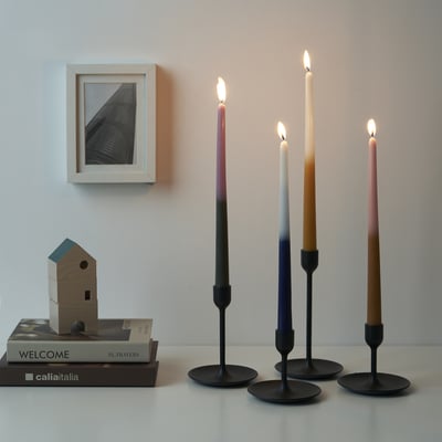 Four lit candles with colorful wicks on a table, beside books and a birdhouse, with a picture above.